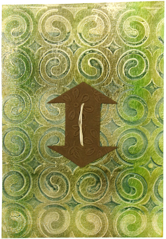 An abstract image with tan and olive green swirls forming circles repeated four across and six down as a background, with a brown textured fabric appliqué in the shape of a double-sided arrow oriented vertically in the center. The arrow is sewn to the page with a piece of raffia.
